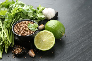 Photo of Coriander seeds in bowl, fresh cilantro, lime and garlic on dark textured table, closeup. Space for text