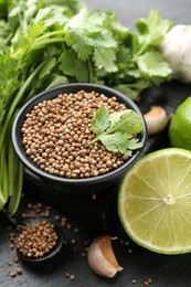Photo of Coriander seeds in bowl, fresh cilantro, lime and garlic on dark textured table, closeup