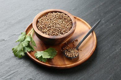 Photo of Coriander seeds in bowl, spoon and fresh cilantro leaves on dark textured table, closeup
