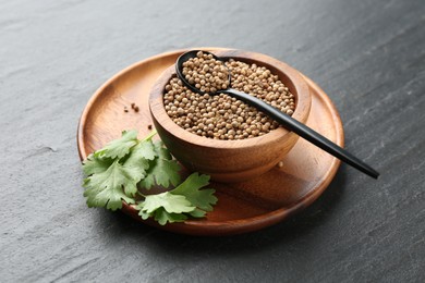 Photo of Coriander seeds in bowl, spoon and fresh cilantro leaves on dark textured table, closeup