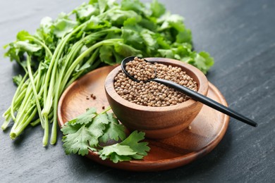 Photo of Coriander seeds in bowl, spoon and fresh cilantro leaves on dark textured table, closeup
