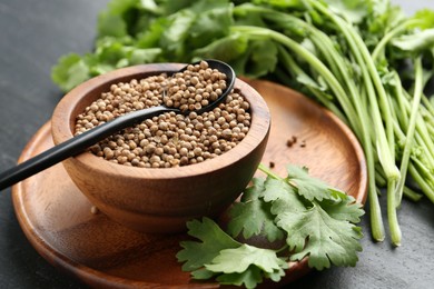 Photo of Coriander seeds in bowl, spoon and fresh cilantro leaves on dark textured table, closeup