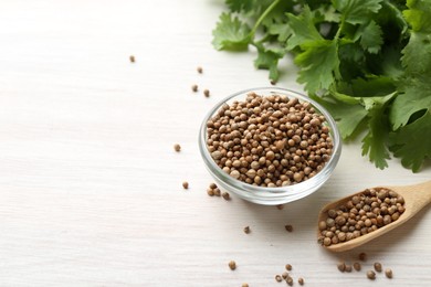 Photo of Coriander seeds in bowl, spoon and fresh cilantro leaves on light wooden table, closeup. Space for text