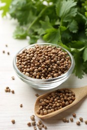 Photo of Coriander seeds in bowl, spoon and fresh cilantro leaves on light wooden table, closeup