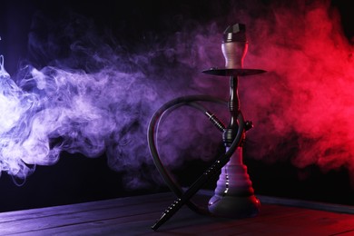 Photo of Hookah on wooden table with smoke in color lights against black background