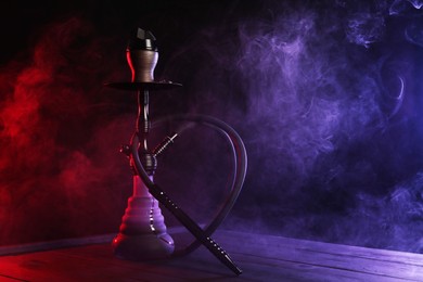 Photo of Hookah on wooden table with smoke in color lights against black background