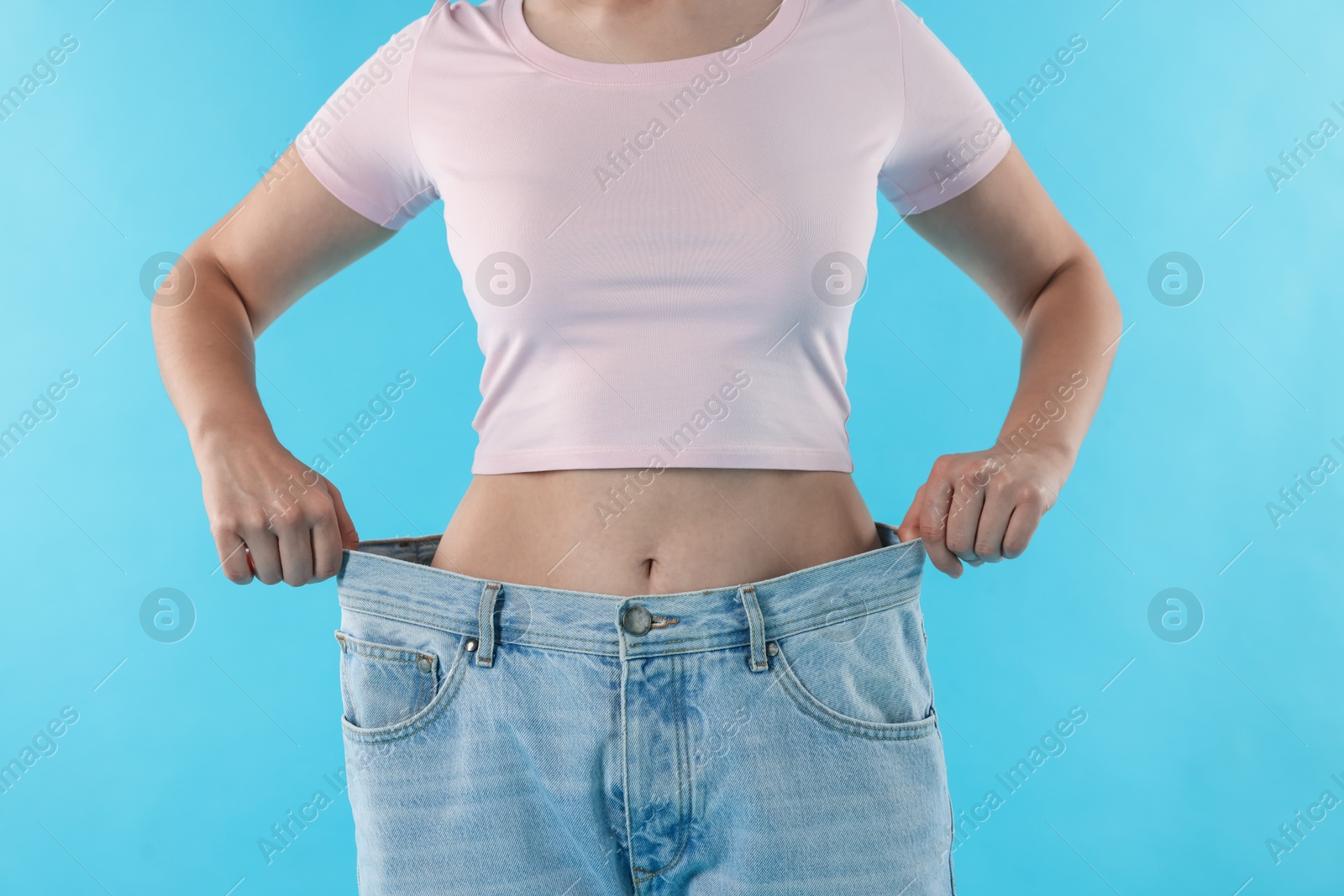 Photo of Woman wearing old jeans after weight loss on light blue background, closeup