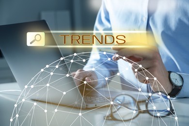 Image of Businessman using laptop and virtual screen to find hot tendencies, new ideas, popular directions at table, closeup. Search bar with word Trends and globe