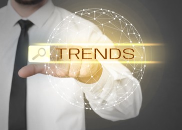 Image of Businessman using virtual screen to find hot tendencies, new ideas, popular directions against grey background, closeup. Search bar with word Trends and globe