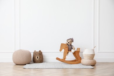 Photo of Rocking horse with bear toy, pouf and wicker baskets near white wall in child room. Interior design