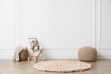 Photo of Toy stroller with bear, pouf and wicker basket near white wall in child room. Interior design