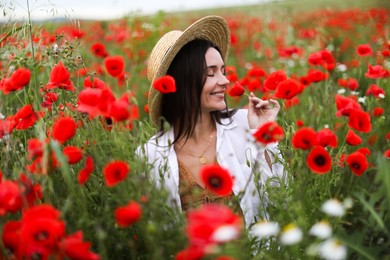 Photo of Smiling woman in hat sniffing red poppy flowers in wildflower meadow
