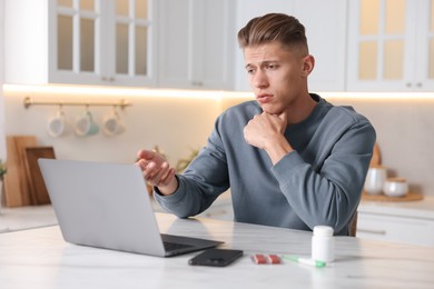 Photo of Sick man having online consultation with doctor via laptop at white marble table in kitchen