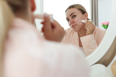 Photo of Woman applying cream onto face near mirror indoors. Acne treatment