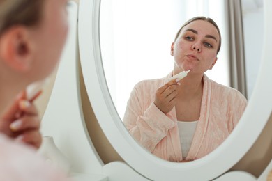 Photo of Woman applying cream onto face near mirror indoors. Acne treatment