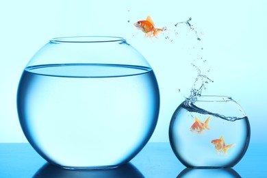 Image of Goldfish jumping out of small glass bowl into bigger one against light blue background