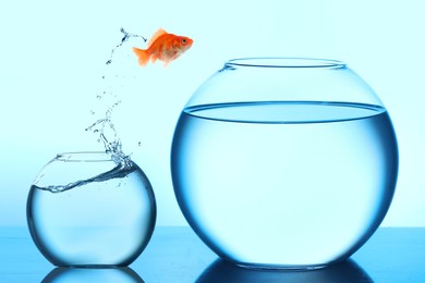 Image of Goldfish jumping out of small glass bowl into bigger one against light blue background