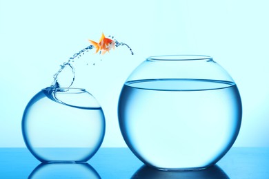Image of Goldfish jumping out of small glass bowl into bigger one against light blue background