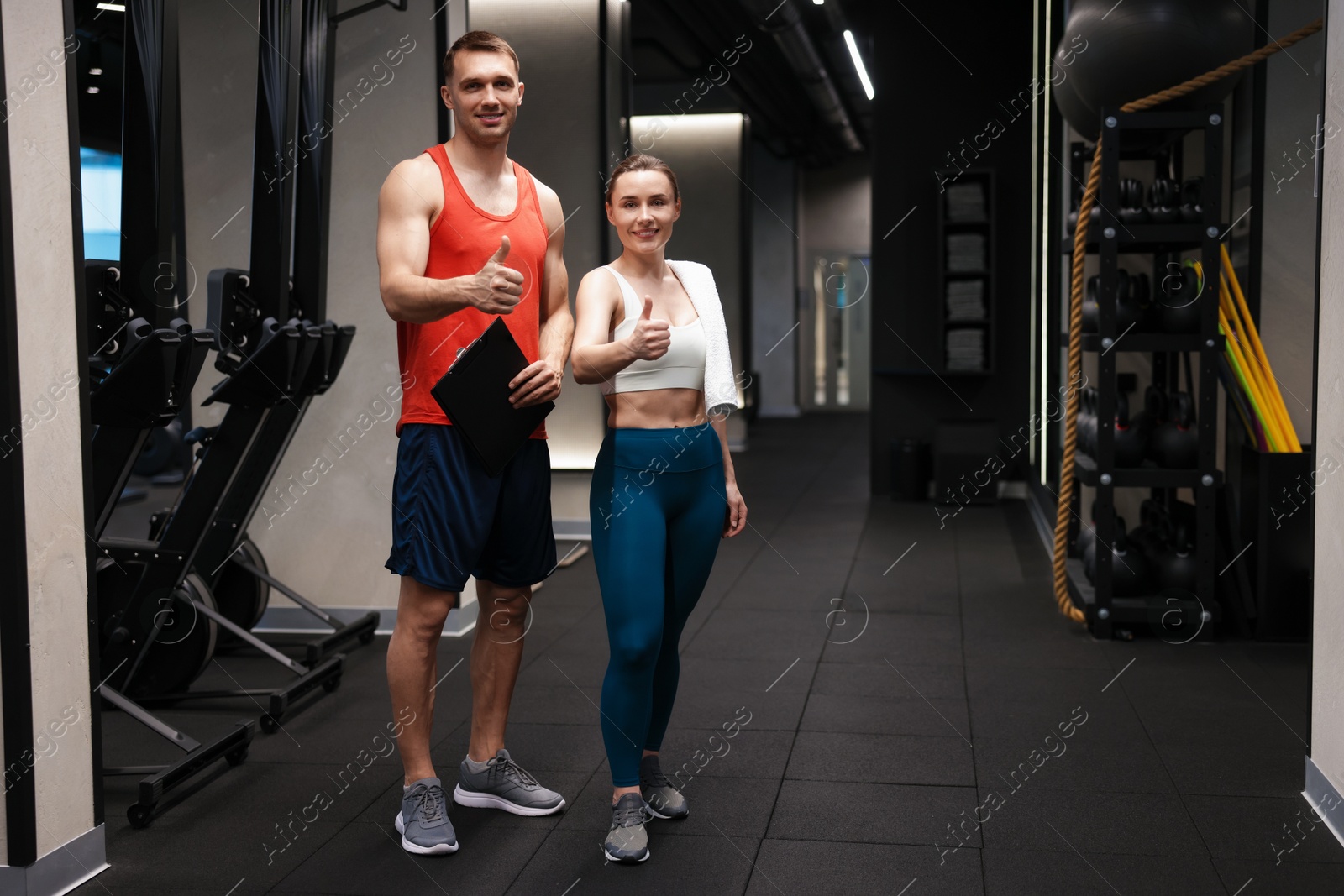Young woman with professional personal trainer showing thumbs up in gym Photo of Young woman with professional personal trainer showing thumbs up in gym