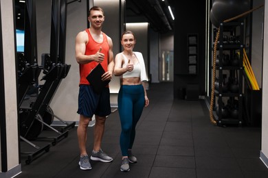 Photo of Young woman with professional personal trainer showing thumbs up in gym