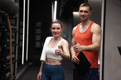 Photo of Young woman with professional personal trainer showing thumbs up in gym