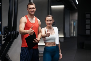Photo of Young woman with professional personal trainer showing thumbs up in gym