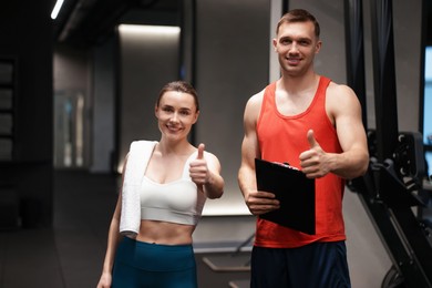 Photo of Young woman with professional personal trainer showing thumbs up in gym