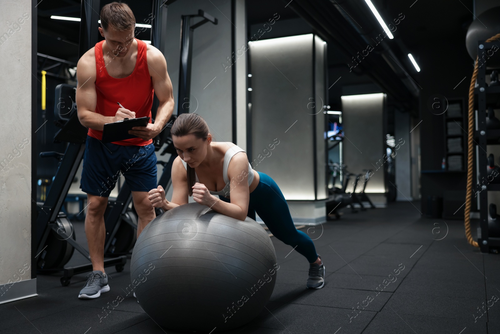 Young woman exercising with fitness ball near professional personal trainer in gym, space for text Photo of Young woman exercising with fitness ball near professional personal trainer in gym, space for text