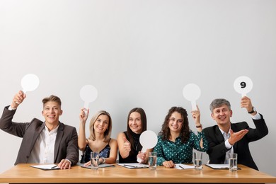 Photo of Panel of happy judges voting with score signs at table against light grey background