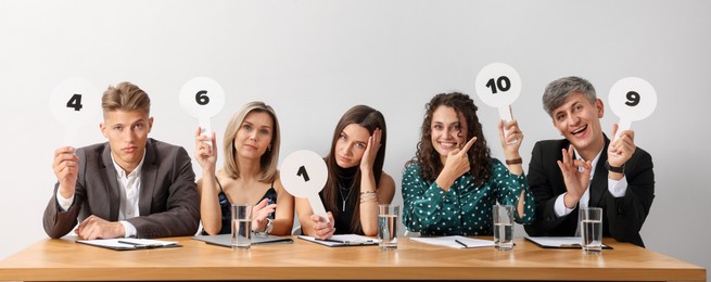 Photo of Panel of emotional judges voting with score signs at table against light grey background