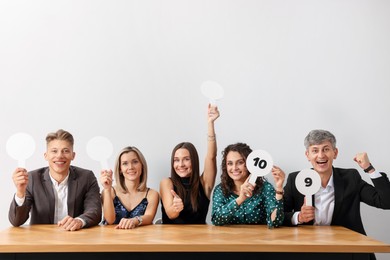 Photo of Panel of happy judges voting with score signs at table against light grey background