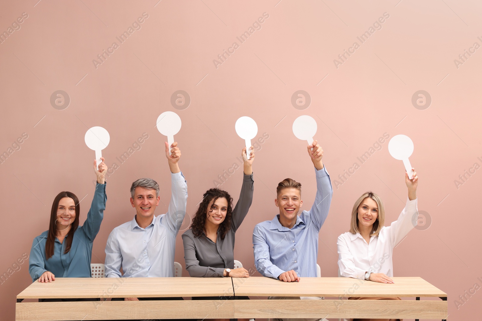 Panel of happy judges voting with blank score signs at tables against beige background Photo of Panel of happy judges voting with blank score signs at tables against beige background
