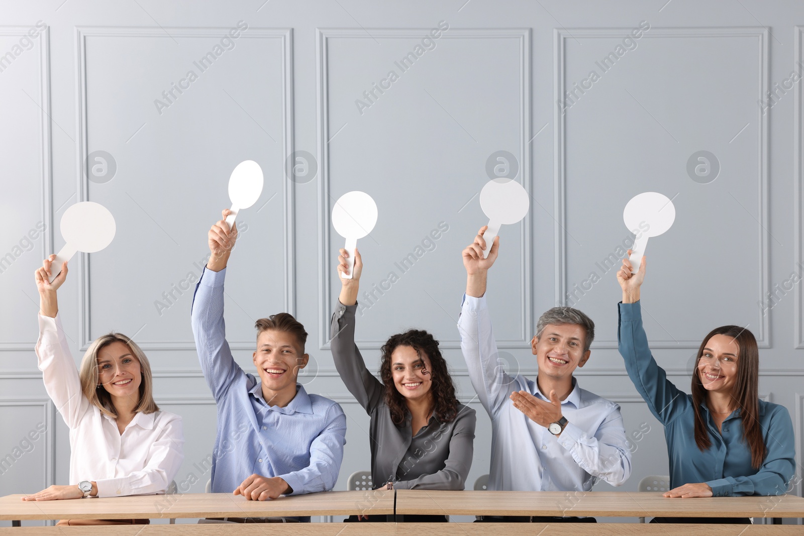 Panel of happy judges voting with blank score signs at tables near grey wall Photo of Panel of happy judges voting with blank score signs at tables near grey wall