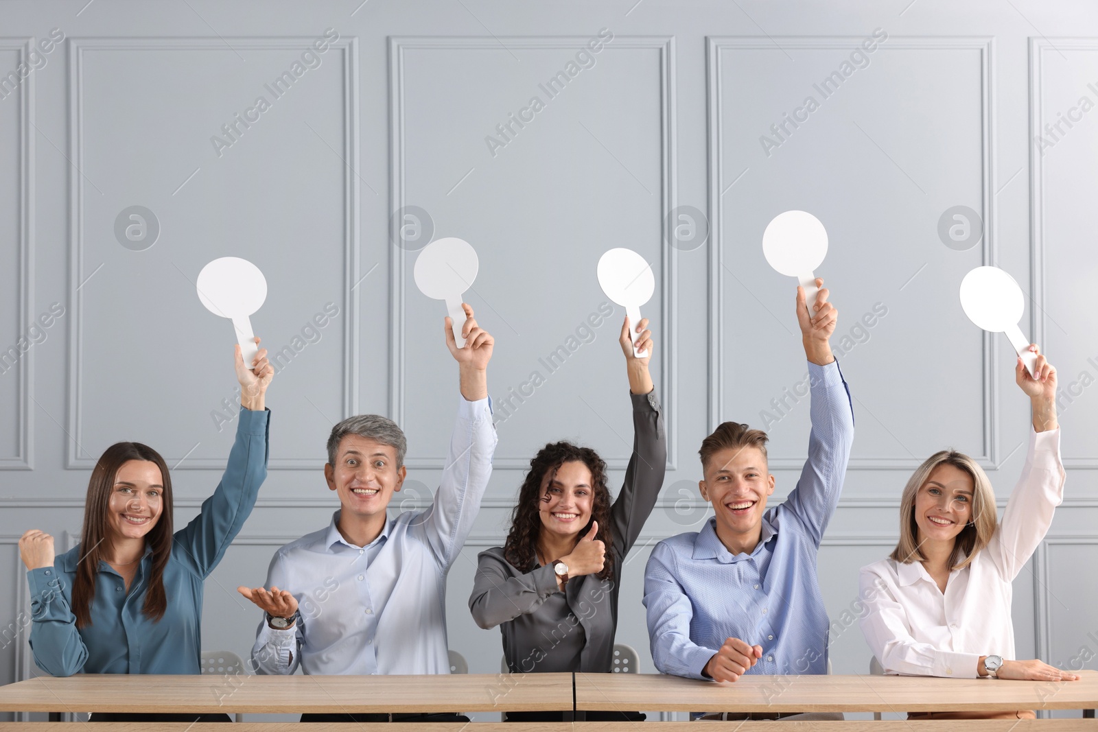Panel of happy judges voting with blank score signs at tables near grey wall Photo of Panel of happy judges voting with blank score signs at tables near grey wall
