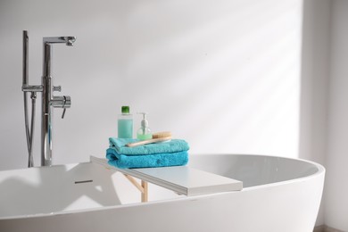Photo of White board with soft towels, brush and cosmetic products on tub indoors