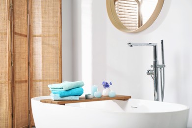 Photo of Wooden board with soft towels, sea salt, flowers and burning candles on tub in bathroom