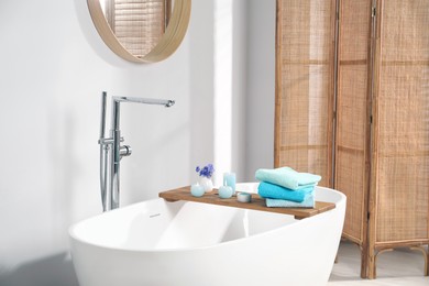 Photo of Wooden board with soft towels, sea salt, flowers and burning candles on tub in bathroom