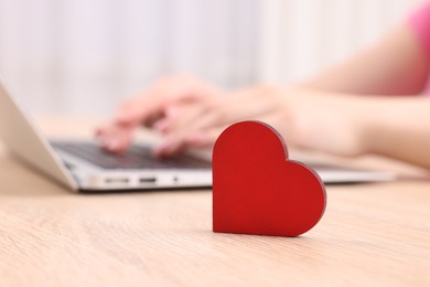 Photo of Online dating site. Woman using laptop at table indoors, focus on decorative heart