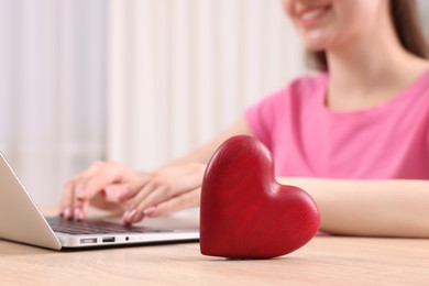 Photo of Online dating site. Woman using laptop at table indoors, focus on decorative heart