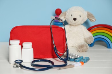 Photo of Pediatrician concept. Toys, first aid kit, pills, thermometer and stethoscope on white wooden table against light blue background, closeup