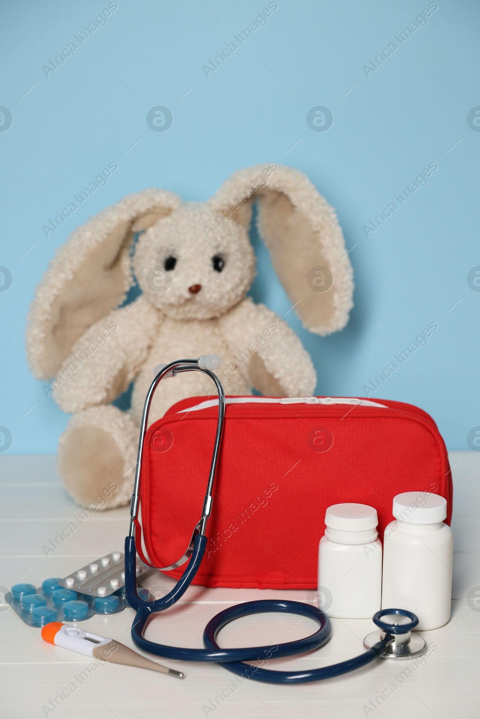 Pediatrician concept. Toy bunny, first aid kit, pills, thermometer and stethoscope on white wooden table against light blue background Photo of Pediatrician concept. Toy bunny, first aid kit, pills, thermometer and stethoscope on white wooden table against light blue background