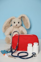Photo of Pediatrician concept. Toy bunny, first aid kit, pills, thermometer and stethoscope on white wooden table against light blue background