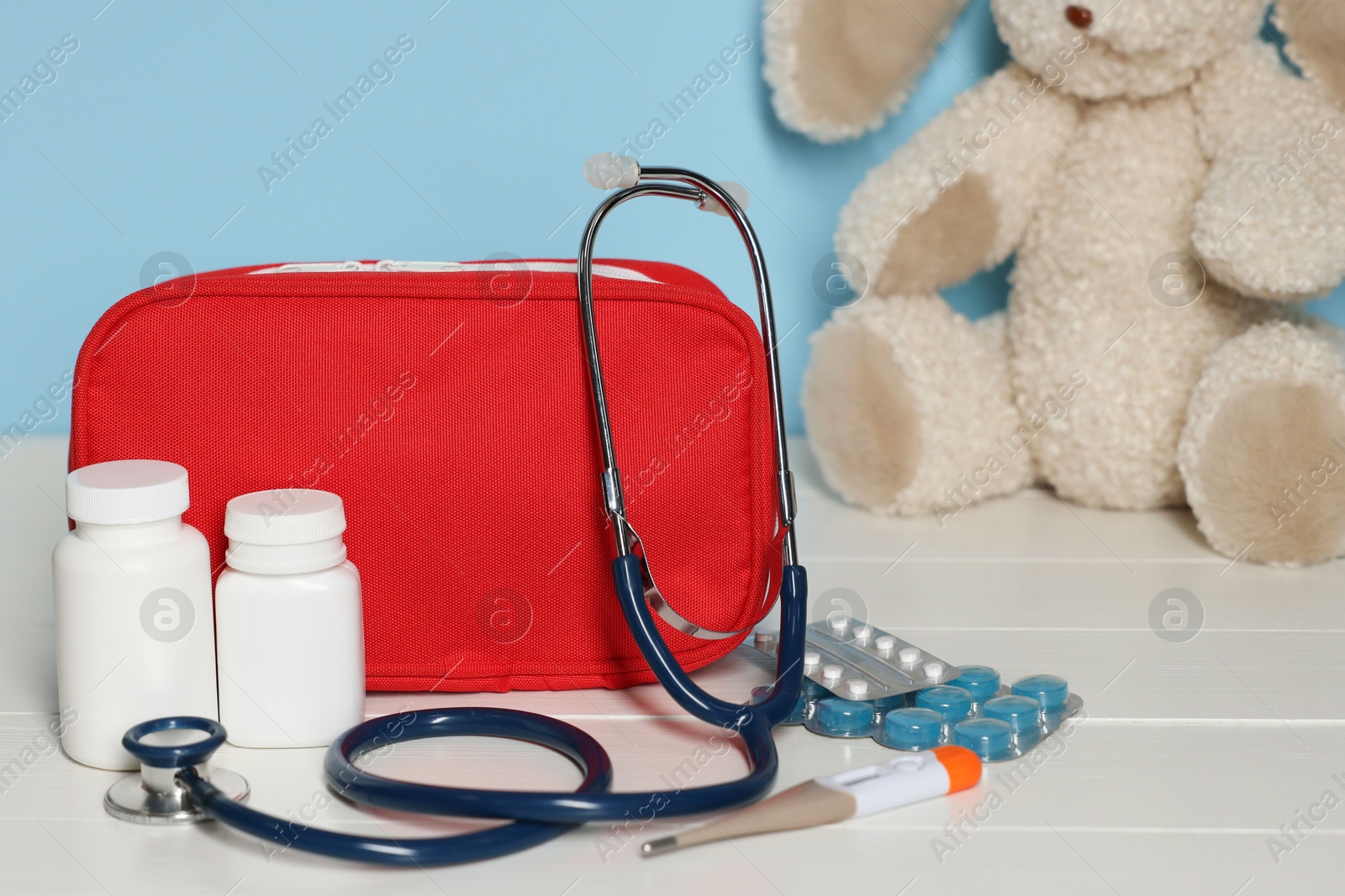 Pediatrician concept. First aid kit, pills, thermometer, stethoscope and toy bunny on white wooden table against light blue background, closeup Photo of Pediatrician concept. First aid kit, pills, thermometer, stethoscope and toy bunny on white wooden table against light blue background, closeup