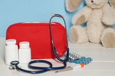 Photo of Pediatrician concept. First aid kit, pills, thermometer, stethoscope and toy bunny on white wooden table against light blue background, closeup