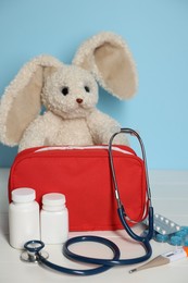 Photo of Pediatrician concept. Toy bunny, first aid kit, pills, thermometer and stethoscope on white wooden table against light blue background, closeup