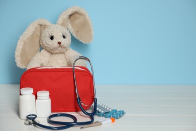 Photo of Pediatrician concept. Toy bunny, first aid kit, pills, thermometer and stethoscope on white wooden table against light blue background, closeup. Space for text