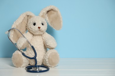 Photo of Pediatrician concept. Toy bunny and stethoscope on white wooden table against light blue background, space for text