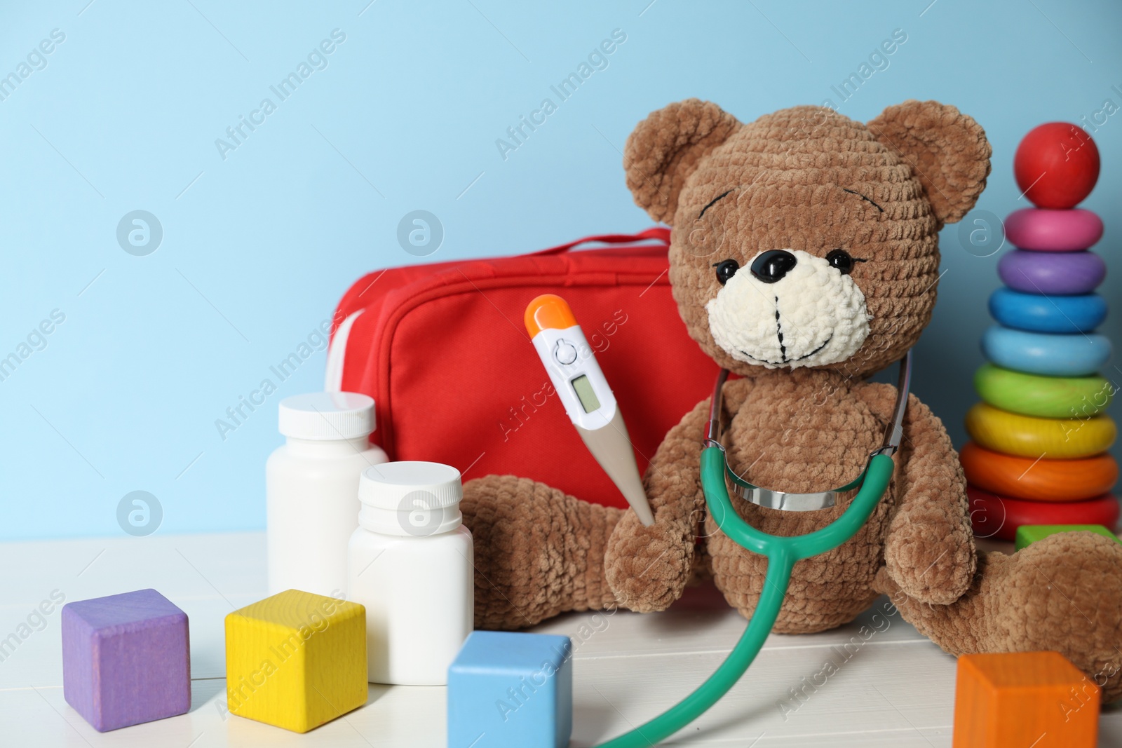 Pediatrician concept. Toys, first aid kit, pills, thermometer and stethoscope on white wooden table against light blue background, closeup Photo of Pediatrician concept. Toys, first aid kit, pills, thermometer and stethoscope on white wooden table against light blue background, closeup