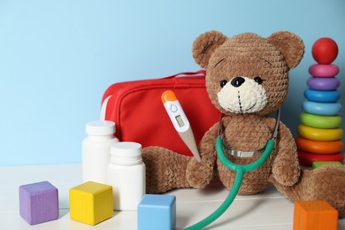 Photo of Pediatrician concept. Toys, first aid kit, pills, thermometer and stethoscope on white wooden table against light blue background, closeup