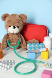 Photo of Pediatrician concept. Toy bear, cubes, first aid kit, pills, thermometer and stethoscope on white wooden table against light blue background, closeup